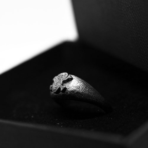 Ανδρικό δαχτυλίδι από ασήμι 925 βαθμών που έχει δεχθεί μαύρη επιπλατίνωση. Το δαχτυλίδι φέρει ένθετο έντεχνο χαρακτικό σχέδιο με νεκροκεφαλή. Δημιουργία της συλλογής ''Θυμήσου Σώμα''